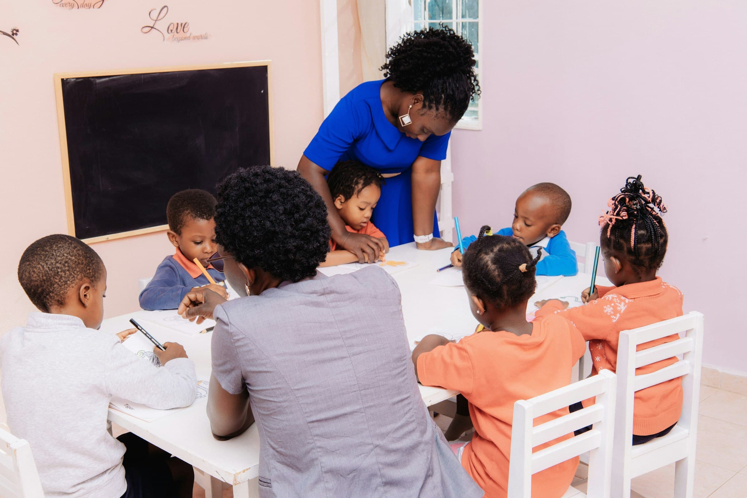 Two adults working alongside a group of young children drawing at a low white table