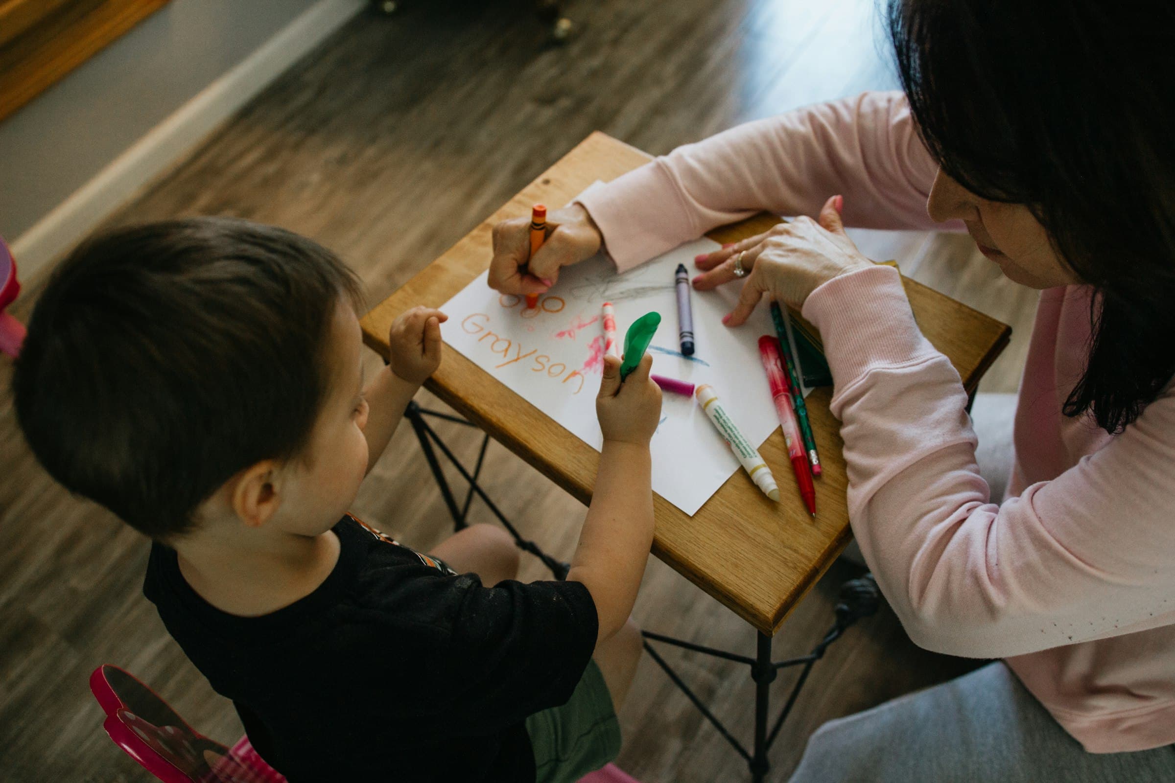 An adult and a young child drawing together with crayons and markers at a small wooden table