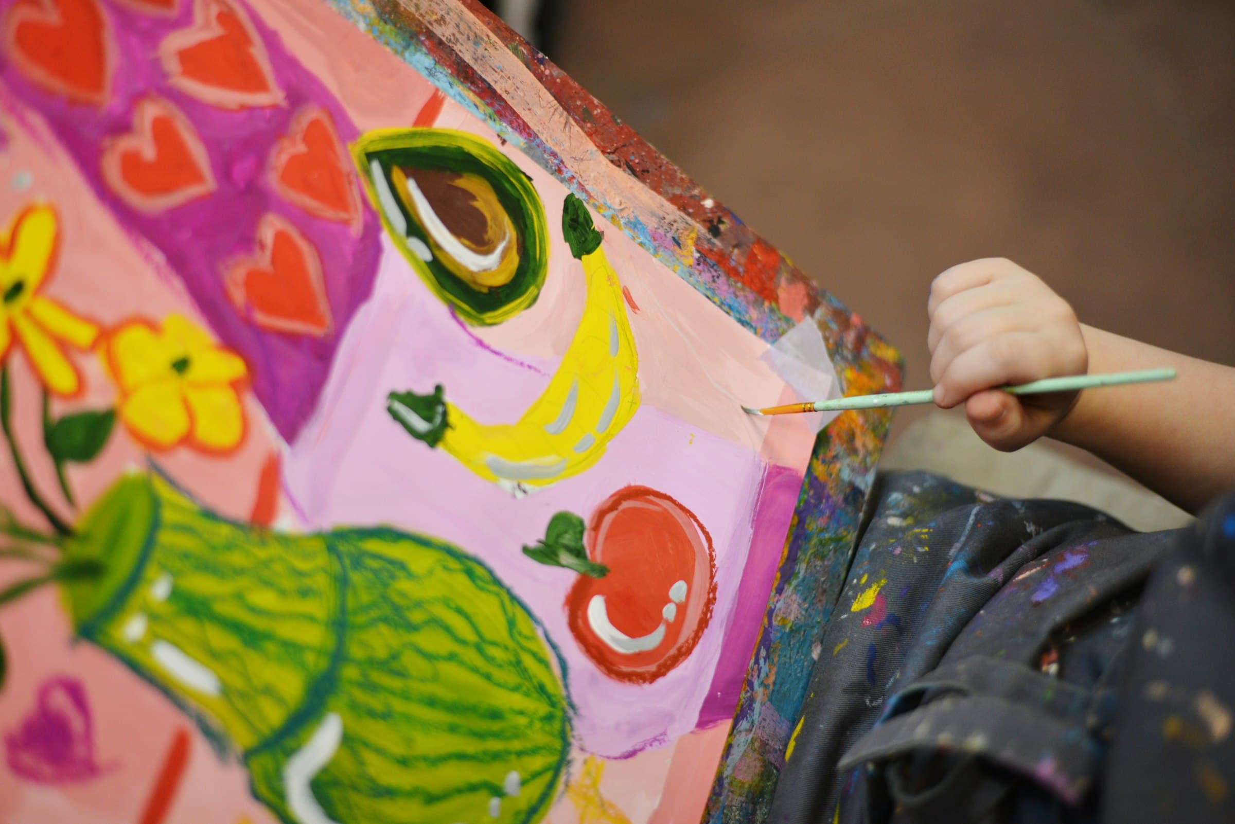 A child's hand guiding a fine brush across a large canvas painted with bright flowers and fruit