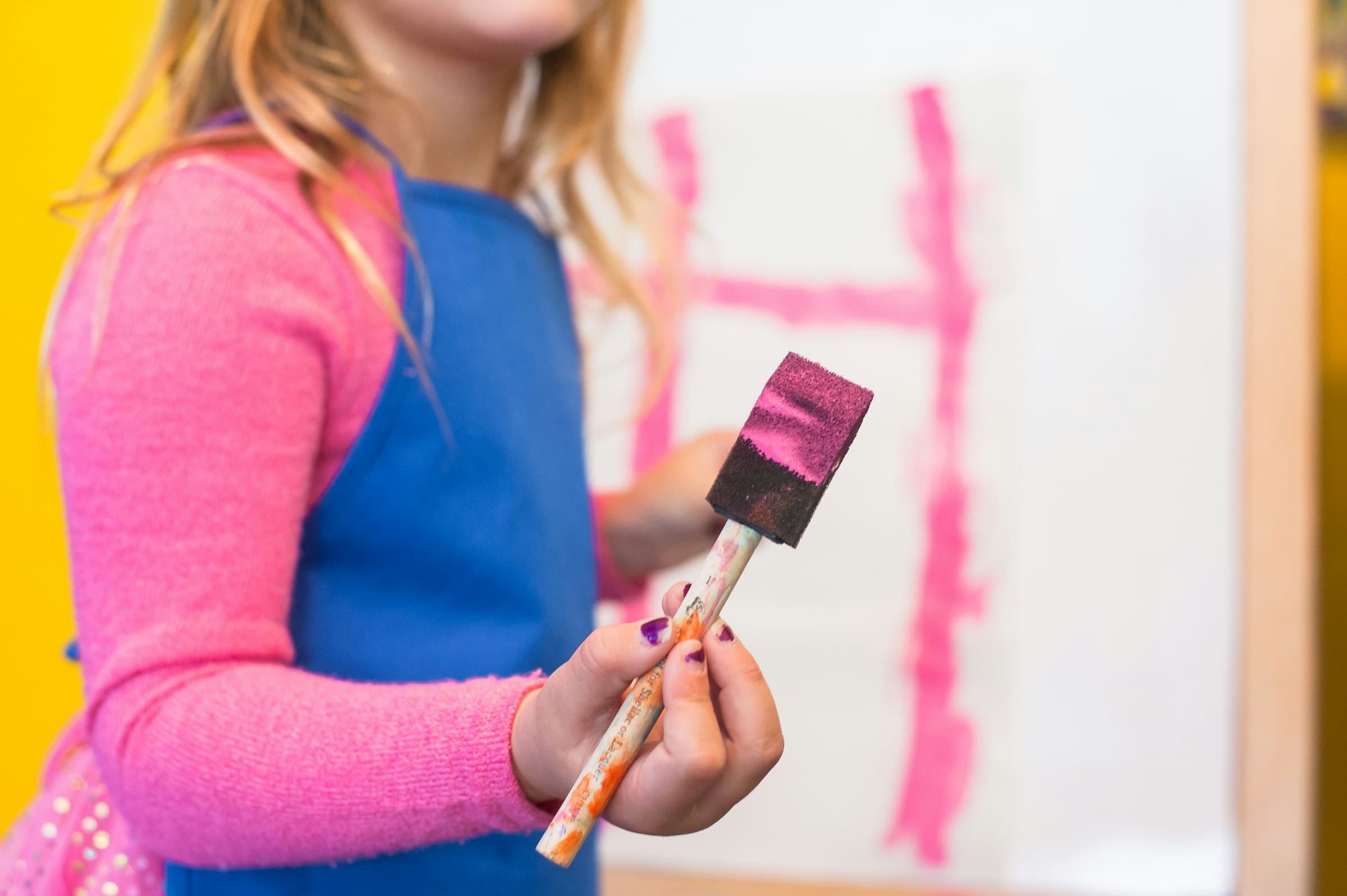 A child in a blue art smock holding a foam paintbrush in front of an easel marked with pink brushstrokes