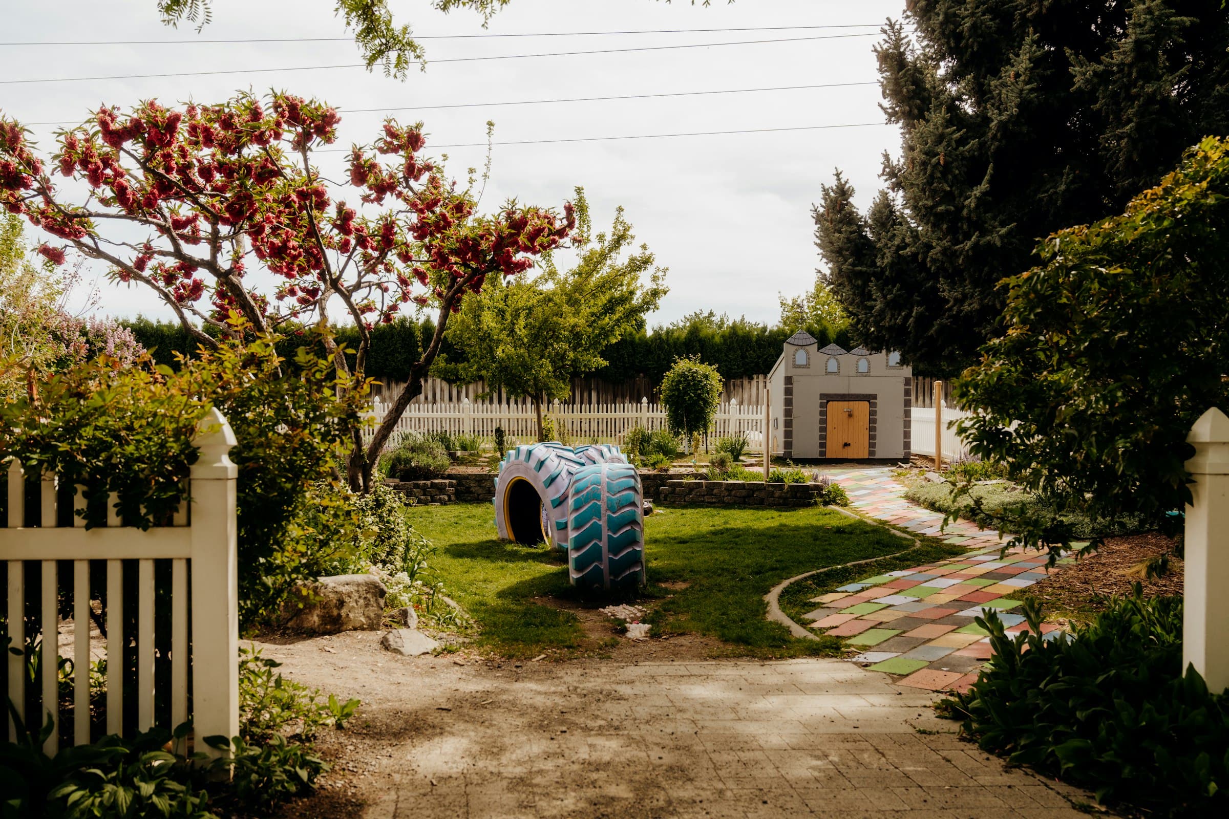 A garden lawn with a blossoming tree, a white picket fence, a painted tyre to climb through, and a path of coloured tiles leading to a small playhouse