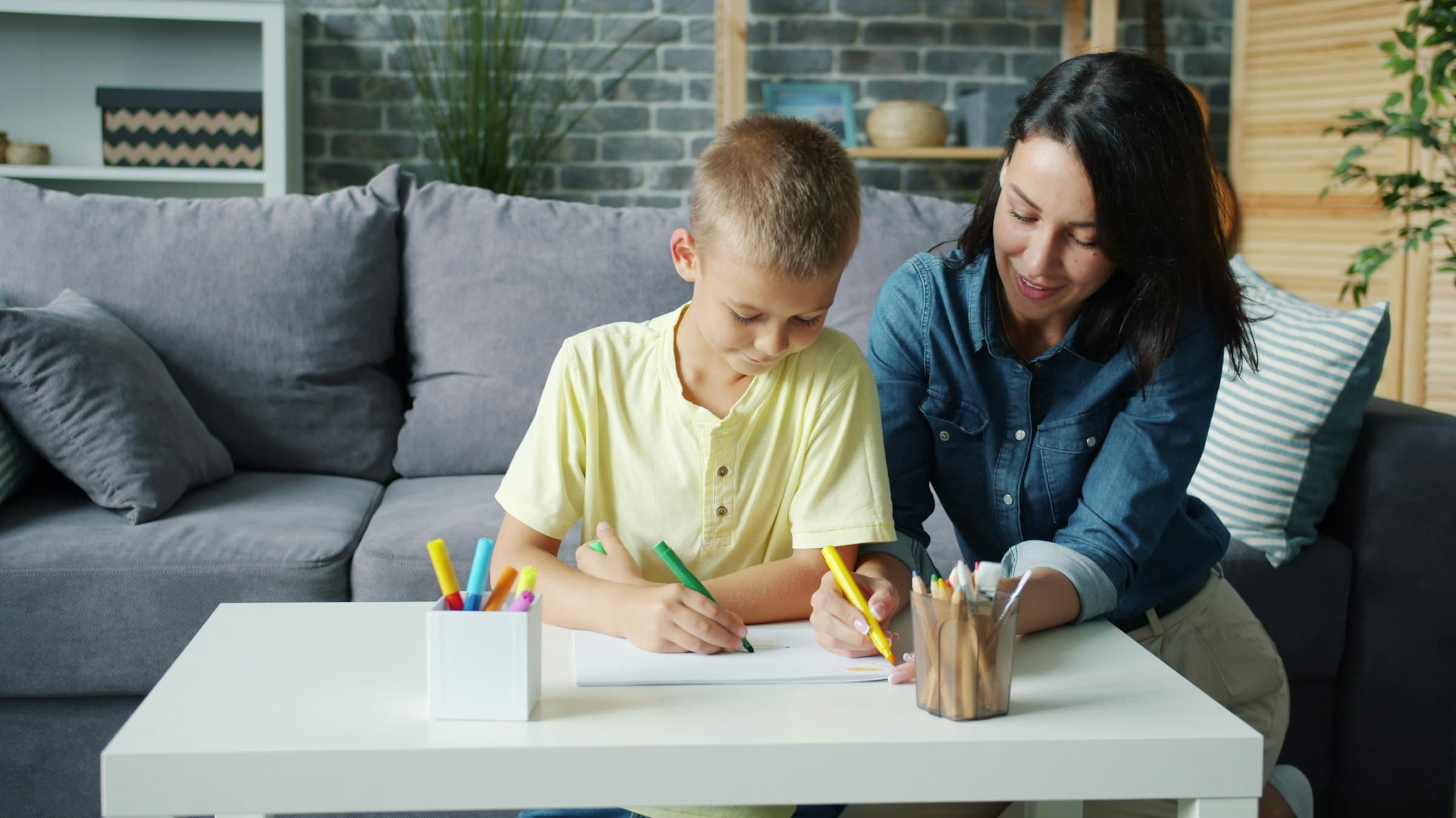 A boy in a yellow t-shirt drawing with a green marker at a low living-room table while a woman beside him leans in to watch