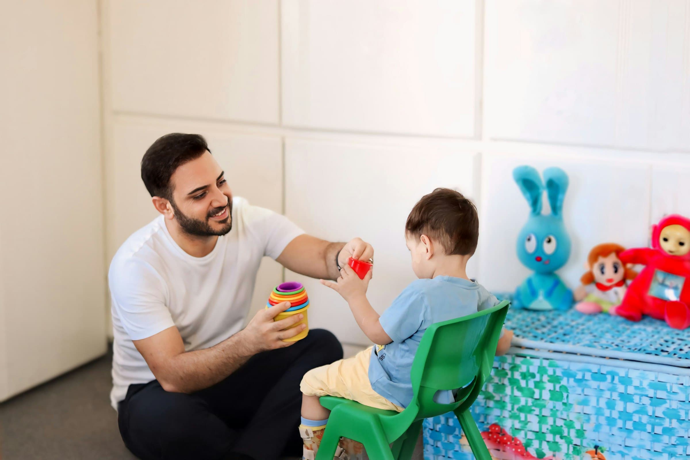An adult sitting on the floor offering a orange stacking cup to a young child on a small green chair
